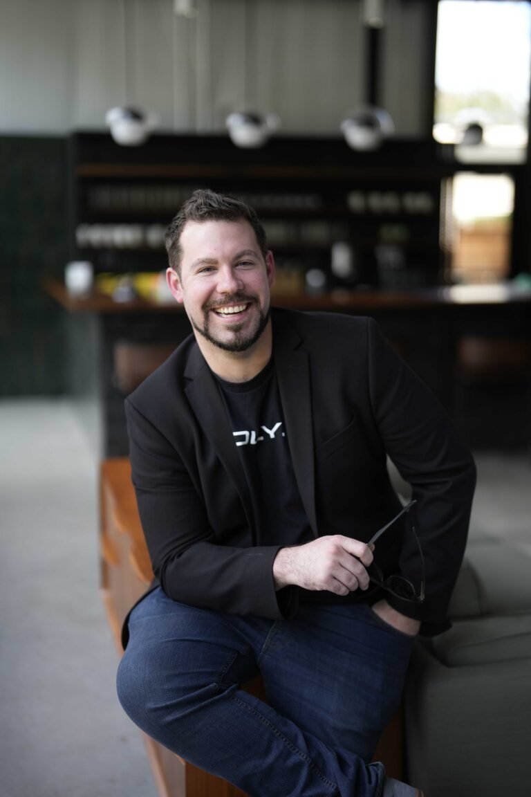 Pat Brennan CEO of Cadly Smiling man wearing a black blazer and a casual shirt, sitting on a wooden bench in a modern interior space, with a blurred background featuring a bar area.