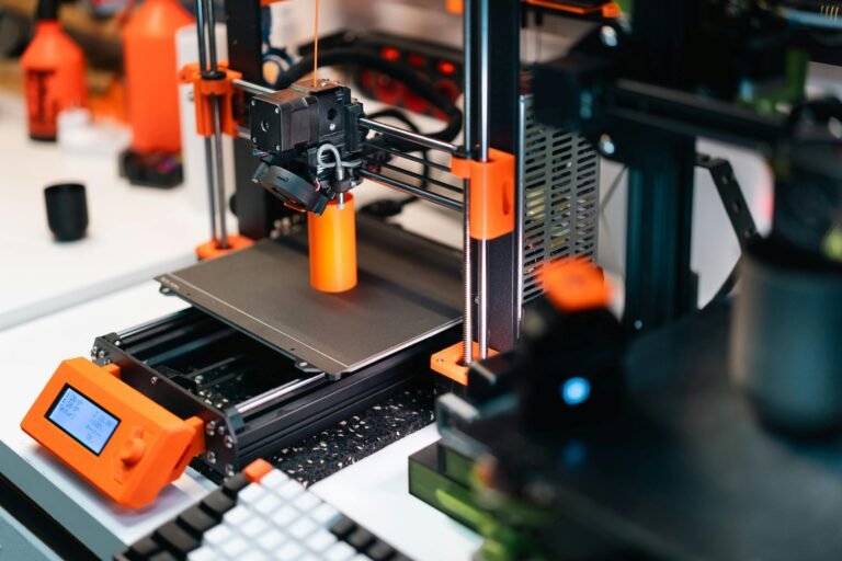 Close-up of a 3D printer in operation, showcasing the hotend and filament extruding an orange cylindrical object. The printer features orange accents, a digital control panel, and a build plate, set in a modern workspace with various tools and materials in the background.
