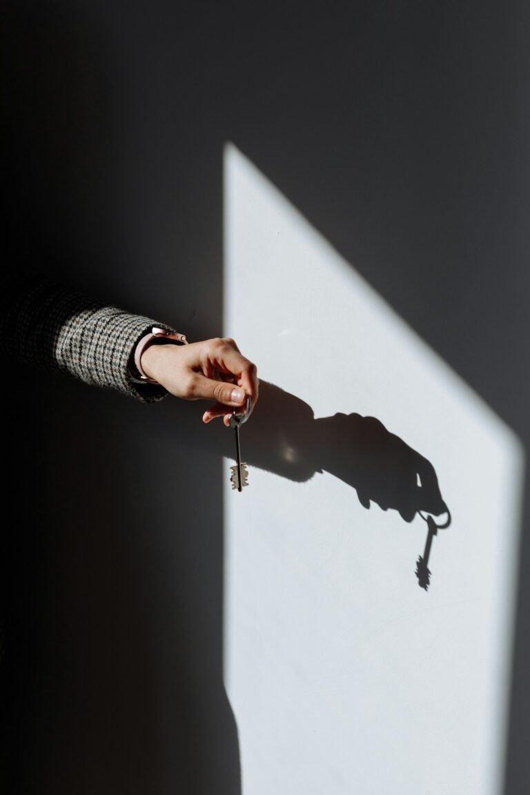 A hand holding a set of keys casts a shadow on a wall, with sunlight creating a dramatic contrast between light and dark areas.