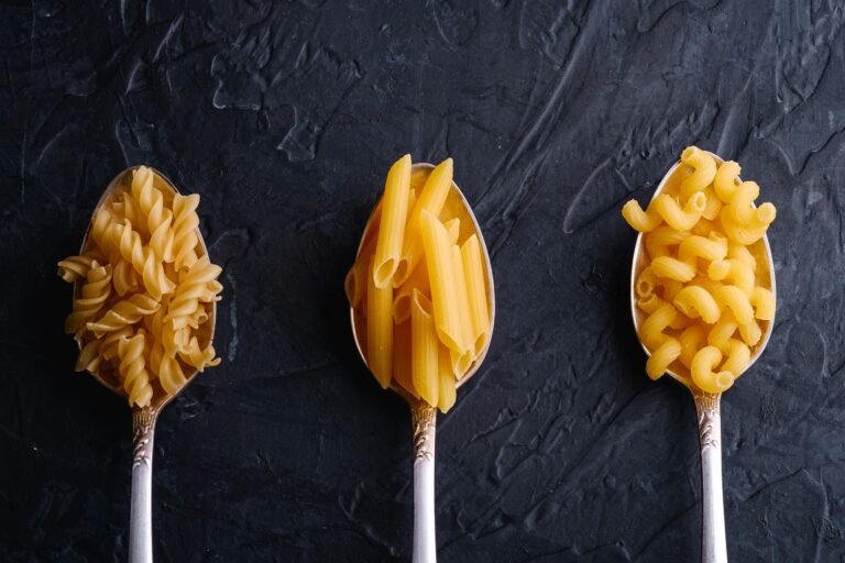 Three types of uncooked pasta displayed in vintage silver spoons, featuring fusilli, penne, and elbow macaroni, against a dark textured background. Ideal for showcasing pasta varieties for recipes or cooking blogs.