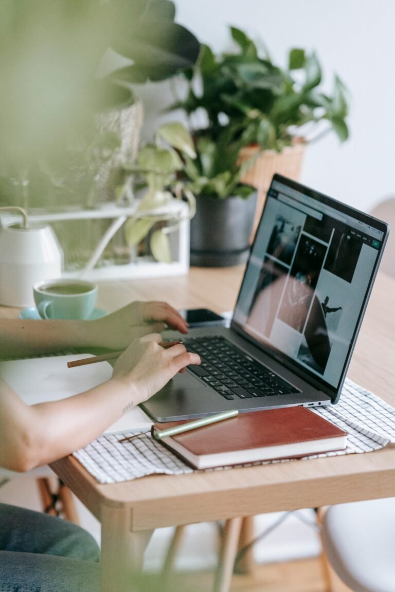 Person working on a laptop at a wooden table, surrounded by plants, a cup of coffee, and notebooks. The workspace features a modern aesthetic with a focus on productivity and creativity.