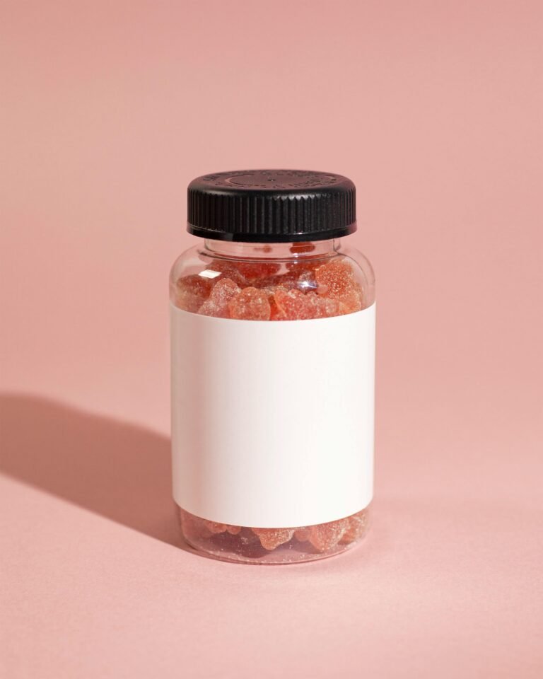 Clear plastic bottle filled with colorful gummy vitamins, featuring a black screw-on cap and a blank white label, set against a soft pink background.